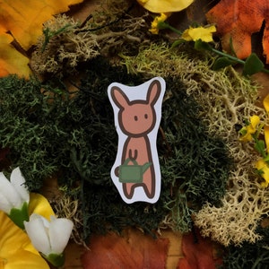 May include: A brown bunny sticker with a green watering can, set against a background of fall foliage and moss.