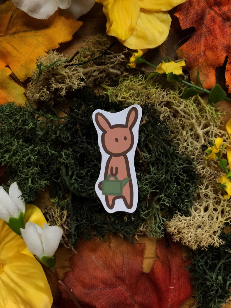 May include: A brown bunny sticker with a green watering can, set against a background of fall foliage and moss.