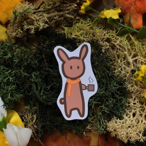 May include: A brown cartoon bunny wearing an orange scarf holds a steaming cup of coffee. The bunny is on a white background with a fall-themed background of moss and leaves.