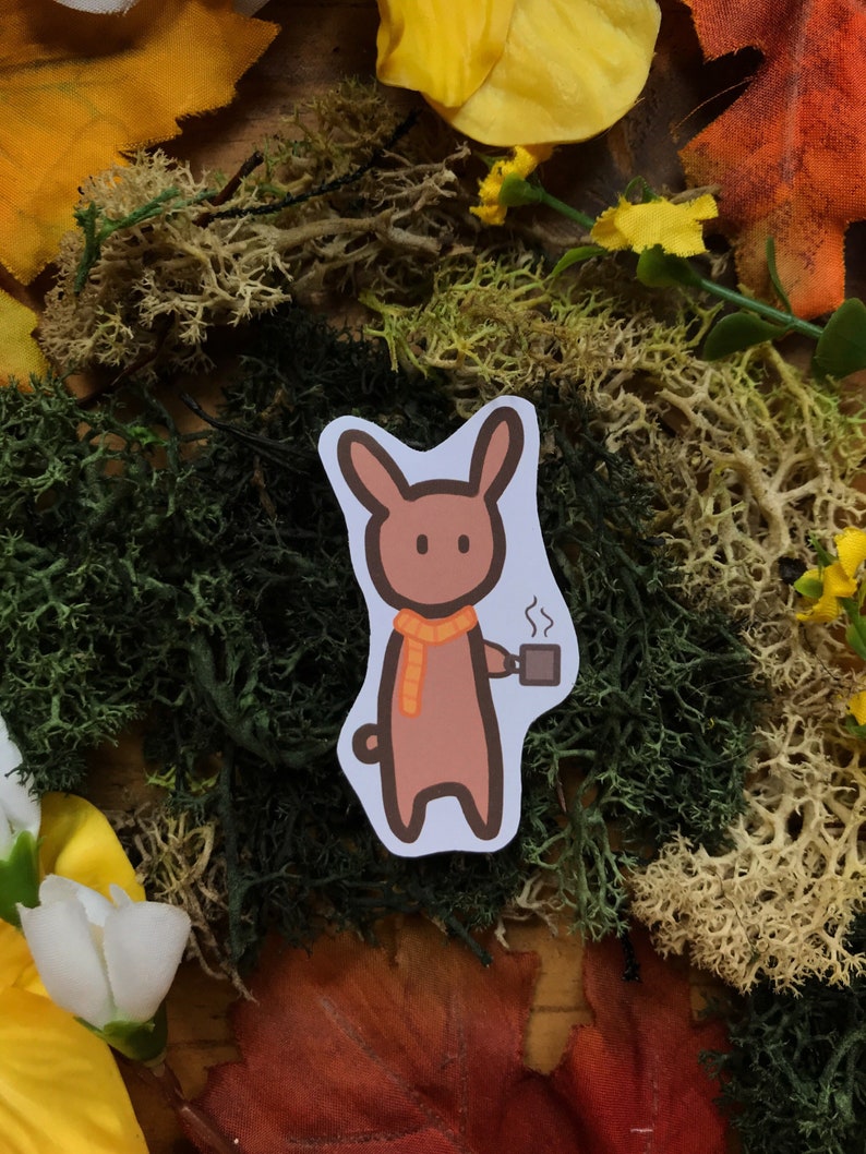 May include: A brown cartoon bunny wearing an orange scarf holds a steaming cup of coffee. The bunny is on a white background with a fall-themed background of moss and leaves.
