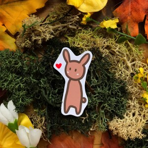 May include: A brown cartoon bunny sticker with a red heart above its head. The sticker is on a background of green moss, yellow flowers, and brown leaves.