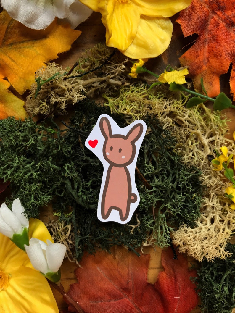 May include: A brown cartoon bunny sticker with a red heart above its head. The sticker is on a background of green moss, yellow flowers, and brown leaves.