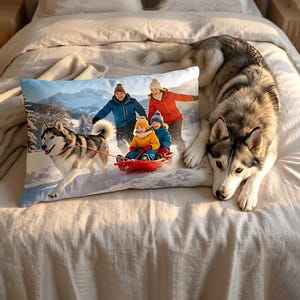 May include: A rectangular pillow featuring a winter scene with a family sledding, pulled by a husky dog. The family is wearing winter clothing. A real husky dog is lying next to the pillow on a bed.