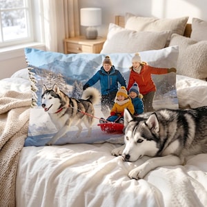 May include: A rectangular pillow with a winter scene featuring a family sledding, pulled by a husky dog. Another husky dog rests on the bed. The pillow has a blue and white mountain backdrop.