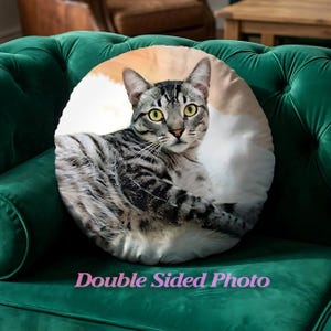 May include: Circular cushion with a photographic print of a grey tabby cat with yellow eyes. The cushion has a white and tan background. Text on the cushion reads "Double Sided Photo". The cushion is on a green velvet sofa.
