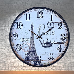 May include: A round wall clock with a black metal frame. The clock face is white with black numerals and hands. It features an illustration of the Eiffel Tower, the word "Paris," and the year "1889."