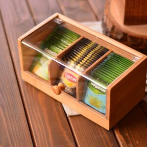 May include: A wooden tea box with a clear lid, containing various tea bags. The box has three compartments, each filled with different types of tea. The box is made of light-colored wood and has a small wooden knob on the front.