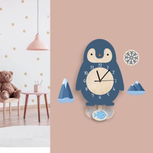 Puede incluir: Un reloj de pared azul en forma de pingüino con una cara de madera y manecillas negras. El reloj tiene un copo de nieve blanco y dos montañas azules con cumbres blancas de nieve a cada lado. El reloj está colgado en una pared rosa.