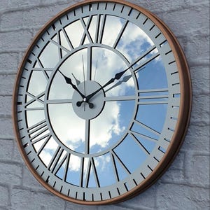 May include: A large, round wall clock with a copper-coloured frame and a mirrored face reflecting a blue sky with clouds. The clock features black hands and Roman numeral hour markers. The clock's diameter is approximately 61 cm.
