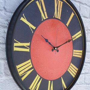 May include: A round wall clock with a black frame and a navy blue face. The clock features gold Roman numerals and black clock hands. The center of the clock is a reddish-orange color.
