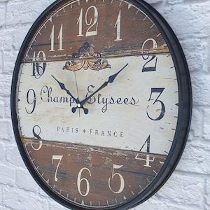 May include: A round wall clock with a black metal frame. The clock face features a distressed wood-look design with large white numerals and the words "Champs Elysees PARIS + FRANCE". The clock hands are black.