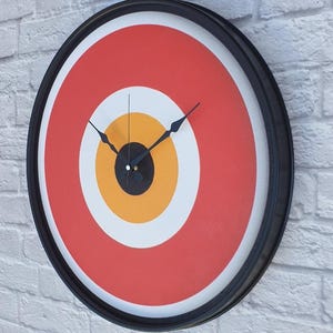 May include: A round wall clock with a black frame. The clock face features a target design with concentric circles in red, white, yellow, and black. The clock hands are black.
