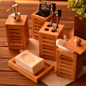 May include: A set of four bamboo bathroom organizers. The set includes a soap dish, a toothbrush holder, a dispenser for liquid soap, and a container with a lid. The organizers are all made of natural bamboo and have a light brown color.