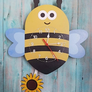 May include: A yellow and black bee-shaped clock with a sunflower pendulum. The clock face has black numbers and a red hour hand and a white minute hand.