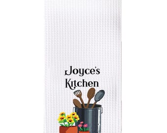 May include: White kitchen towel with a waffle weave texture. The towel features a design of a pot with kitchen utensils and two potted plants. The text "Joyce's Kitchen" is printed in black on the towel.