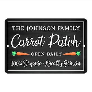 May include: Black and white sign with the text "The Johnson Family Carrot Patch Open Daily 100% Organic Locally Grown" with two orange carrots with green tops on either side of the text.