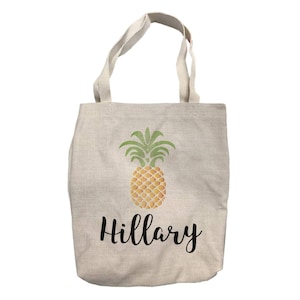 May include: A beige canvas tote bag with a pineapple graphic and the name "Hillary" printed in black.