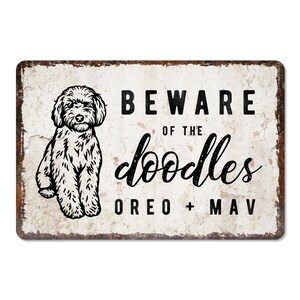 May include: A white metal sign with a distressed look featuring a black and white doodle dog illustration. The sign reads "Beware of the doodles Oreo + Mav".
