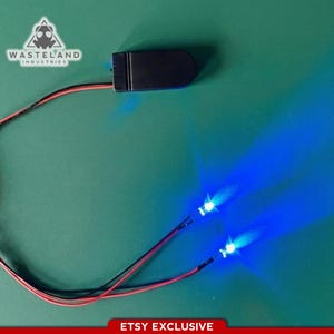 Op de afbeelding: Een zwart batterijpakket met twee blauwe LED-lampjes. De lampjes zijn verbonden door rode en zwarte draden. Het logo van Wasteland Industries staat in de linkerbovenhoek. De tekst "ETSY EXCLUSIVE" staat onderaan.