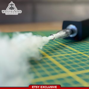 Könnte beinhalten: Ein schwarzes Handgerät mit einer Metalldüse, aus der eine dicke weiße Rauchwolke austritt. Das Gerät trägt das Logo und den Schriftzug Wasteland Industries. Der Gegenstand liegt auf einer grünen Schneidematte mit gelben Linien und einem Etsy Exclusive-Etikett.