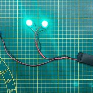 May include: Two bright green LED lights connected by wires to a black battery holder. A blue LED light is also connected to the wires. The components are on a green grid surface.