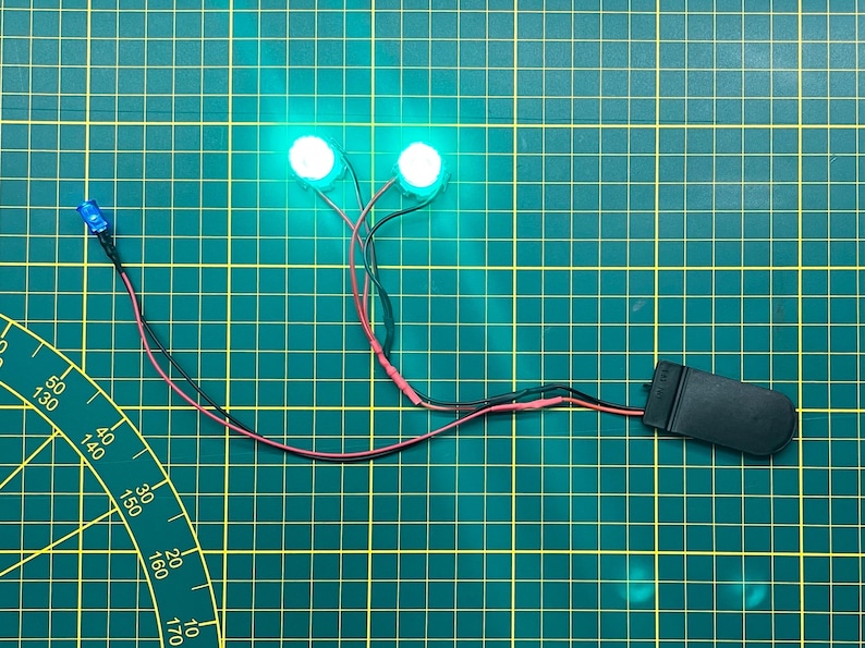May include: Two bright green LED lights connected by wires to a black battery holder. A blue LED light is also connected to the wires. The components are on a green grid surface.