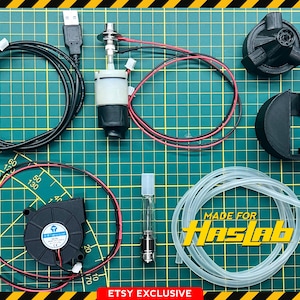 May include: An assortment of black and clear components, including wires, a fan, and a clear tube, arranged on a green grid surface. The Ghostbusters logo is in the upper left corner. Text on the image reads "MADE FOR HASLAB" and "ETSY EXCLUSIVE".