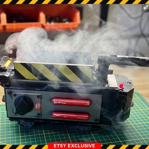 May include: A prop replica of a Ghostbusters proton pack, featuring a black and gray body with yellow and black hazard stripes. Red accents and a handle are visible, with a wisp of smoke emanating from the top. The Ghostbusters logo is in the upper left corner.
