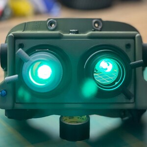 May include: A military green night vision device with two bright green lenses. The device has several knobs and adjustments. It is mounted on a surface with tape.