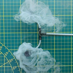 Peut inclure: Gros plan sur un tube en plastique transparent avec deux connecteurs noirs et une valve noire. Le tube est relié à une machine à fumée blanche qui produit une grande quantité de fumée blanche. La fumée tourbillonne autour du tube et des connecteurs. L'arrière-plan est un tapis de coupe vert avec des lignes jaunes.
