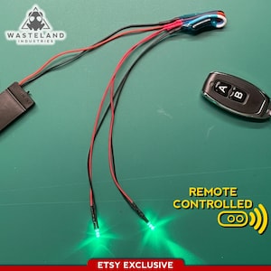 以下が含まれることがあります： 緑色のLEDライト、黒と赤のワイヤー、黒色のバッテリーパックを備えたリモコンデバイス。AとBのボタンが付いたシルバーとブラックのリモコンも含まれています。「Remote Controlled」と「Etsy Exclusive」の文字が見えます。