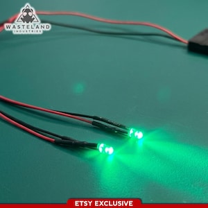 May include: Two small green LED lights with black and red wires, glowing brightly. The wires connect to a black component. The image features the text "WASTELAND INDUSTRIES" and "ETSY EXCLUSIVE" on a teal background.