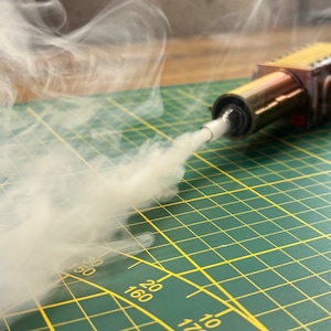 Puede incluir: Primer plano de un pequeño dispositivo metálico con una boquilla que emite una columna de humo blanco. El dispositivo está sobre una estera de corte verde con marcas amarillas.