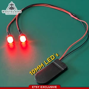 Puede incluir: Dos luces LED rojas de 10 mm conectadas por cables negros y rojos a una caja de baterías negra. El logotipo de Wasteland Industries es visible en la esquina superior izquierda y el texto 10MM LED's está impreso en amarillo cerca de la caja.