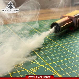 Peut inclure: Un appareil métallique rectangulaire doté d'une buse cylindrique émettant un épais panache de fumée blanche. L'appareil présente une finition cuivrée et un câble noir. Le logo Wasteland Industries est visible dans le coin et le texte Etsy Exclusive apparaît en bas.