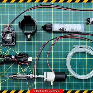 May include: A collection of electronic components, including a black fan, a black bracket, a clear tube, a bottle with a label, and various wires. The items are arranged on a green grid surface with yellow and black borders. The Ghostbusters logo is in the upper left corner.