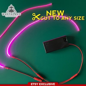 Op de afbeelding: Een neonroze lichtstrip met een zwart batterijpakket en rode draden tegen een groene achtergrond. De tekst "NEW CUT TO ANY SIZE" staat in het geel. Het logo van "Wasteland Industries" staat linksboven. De tekst "ETSY EXCLUSIVE" staat onderaan.