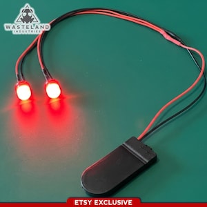 Könnte beinhalten: Zwei leuchtend rote LED-Leuchten, die durch schwarze und rote Drähte mit einem schwarzen Batteriepack verbunden sind. Die Lichter leuchten und werfen einen roten Schein. Das Bild enthält den Text "Wasteland Industries" und "ETSY EXCLUSIVE".