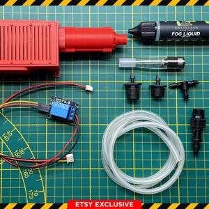 May include: A red prop with a Ghostbusters logo, a black bottle labeled "FOG LIQUID", and various electronic components. The items are arranged on a green grid surface with a yellow and black border. The text "ETSY EXCLUSIVE" is at the bottom.