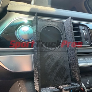 May include: Black textured car phone mount with a circular opening. The mount is positioned in a car's interior, near the dashboard controls. The text "Sport Truck Apps" is visible.