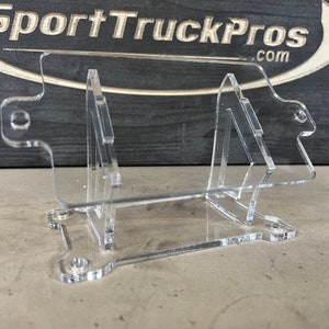 May include: A clear acrylic stand with a rectangular base and two angled supports. The stand is designed to hold a small object, such as a phone or a tablet.