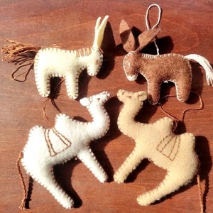 May include: Four handmade felt animal ornaments: a horse, a donkey, and two camels. The horse is off-white with a brown tail, the donkey is brown with a white tail, and the camels are off-white and tan. Each has brown stitched details.