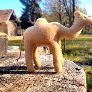 May include: A tan felt camel toy stands on a weathered wooden surface. The handmade camel has a long neck, a hump, and a small tail. The background includes a house and trees, suggesting an outdoor setting.