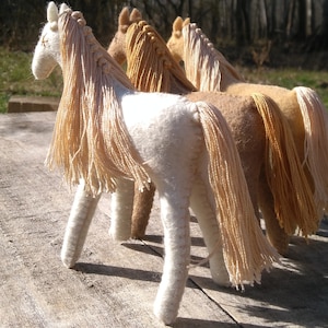 May include: Three handmade felt toy horses with long flowing manes. The horses are white, brown, and tan. The horses are standing on a wooden surface.