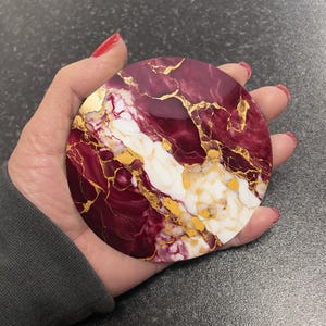 May include: Round resin coaster with a marbled design in shades of burgundy, white, and gold. The coaster features a swirling pattern of burgundy and white with gold accents.