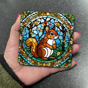 May include: A stained glass coaster featuring a squirrel in a circular frame with a blue, green, and yellow background. The squirrel is brown with a white belly and a bushy tail.