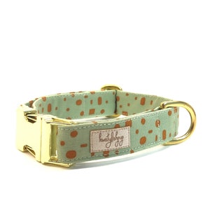 Può includere: Un collare per cani verde con un motivo geometrico marrone e accessori dorati. Il collare ha una fibbia dorata e un anello a D dorato. Il collare è etichettato "kwistylshop".