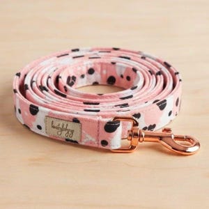 Puede incluir: Una correa para perro enrollada con un patrón de puntos estilo dálmata en rosa y blanco. La correa incluye un mosquetón giratorio de oro rosa metálico y una pequeña etiqueta de tela rectangular con texto en cursiva cosida en el lateral.