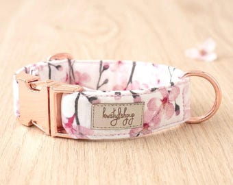 May include: A pet collar featuring a white fabric base with a pink cherry blossom floral pattern. The collar includes a metallic rose gold side-release buckle and a matching D-ring. A rectangular beige fabric label reads kiwistylishpup.