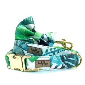 Puede incluir: Un collar para perro verde y blanco con una pajarita a juego con estampado de hojas tropicales. El collar tiene una hebilla dorada y la pajarita tiene un clip dorado. El collar tiene el texto "kwistylshop" en él.